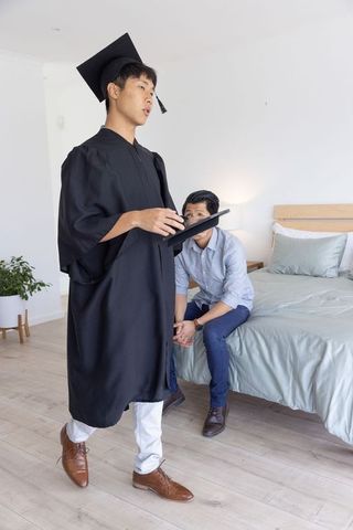 Asian son graduating at home wearing cap and gown holding diploma while father watches