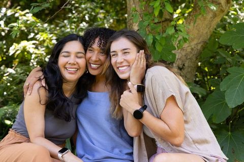 Smiling diverse women hugging and laughing while wearing smartwatches in sunlit park lifestyle