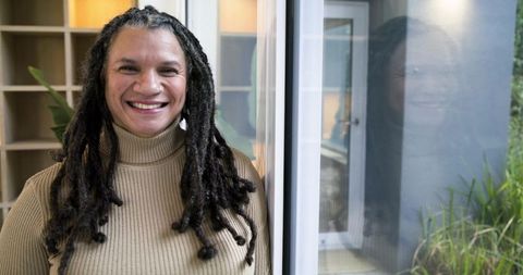 African american woman smiling by window in beige sweater with reflection and cozy interior