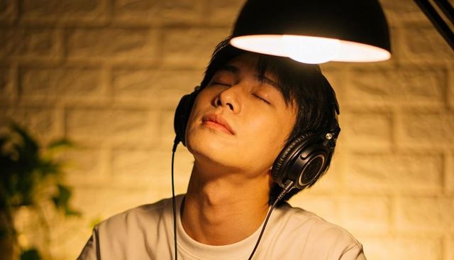 Serene young asian man listening to music with headphones under warm lamp glow in studio