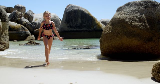 Young girl in a swimsuit walking at a rock-lined beach. Ideal for summer vacation promotions, family holiday advertisements, or coastal lifestyle brochures. Perfect for use in travel blogs, beachwear marketing, and nature-oriented campaigns.