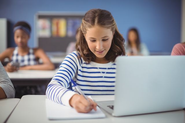 Young girl attentively studying in a classroom setting, using a laptop and writing in a notebook. Ideal for educational content, school promotions, technology in education, and academic resources.