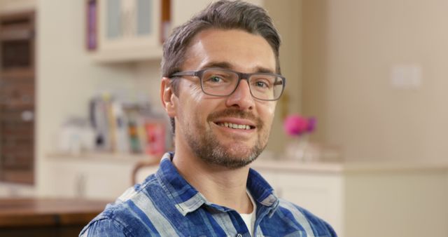 Man smiling while sitting in a cozy living room, wearing glasses and a blue plaid shirt. Ideal for promoting casual fashion, lifestyle blogs, eyewear advertisements, or articles related to home comfort and happiness.