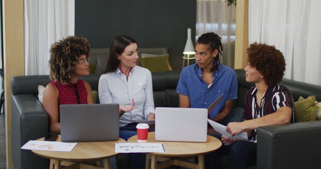 This image shows a diverse group of professionals collaborating and discussing project ideas in a modern office setting. They are seated on couches around a small table, with laptops and documents. Suitable for images promoting teamwork, business culture, office environments, productivity, and technology use in professional settings.