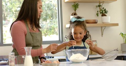 Sunlit kitchen baking: woman guiding child cracking egg into glass mixing bowl