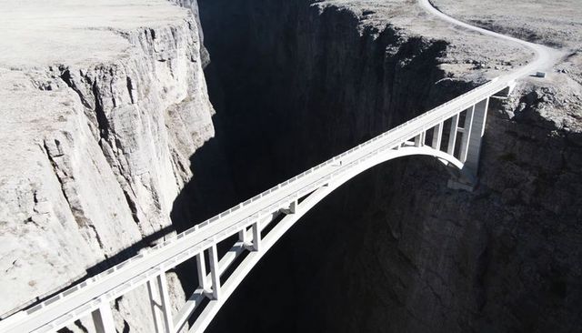 Drone approaching capturing concrete arch bridge spanning deep canyon with winding road