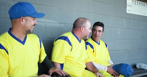 Three baseball teammates laughing and talking in dugout wearing yellow and blue uniforms