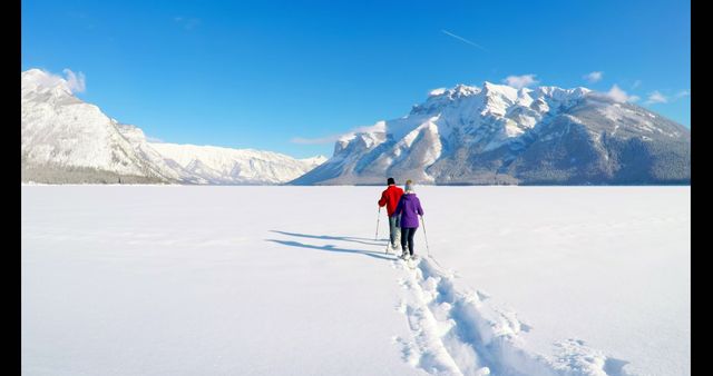Two individuals, one in a red jacket and another in purple, are snowshoeing across a vast snowy terrain with snow-covered mountains in the background. Bright blue sky enhances the crisp, wintry atmosphere. Great for promoting outdoor winter activities, travel, and adventure tourism.