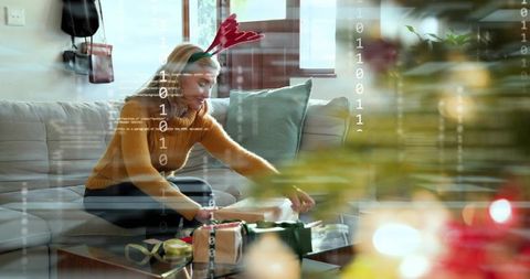 Woman wrapping holiday gifts on sofa wearing antler headband with binary code overlay