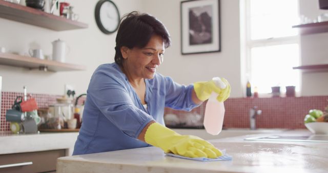 Smiling senior biracial woman wearing gloves and cleaning table in kitchen alone. healthy and active retirement at home.