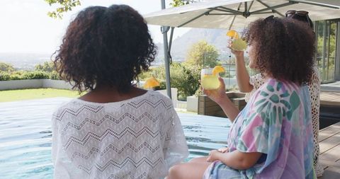 Three women of african descent sipping orange cocktails poolside wearing summer kaftans