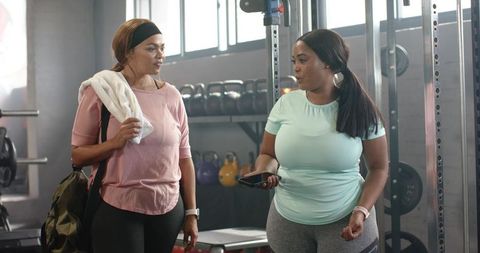 Diverse women chatting after workout in gym holding smartphone and towel wearing leggings