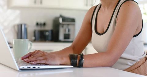 Indian woman typing on laptop at modern kitchen counter with green mug and bracelets