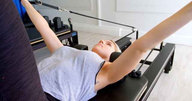 Woman is practicing Pilates on a reformer machine in a bright, indoor space. Ideal for illustrating fitness routines, promoting active lifestyle, or showcasing Pilates exercises and equipment.