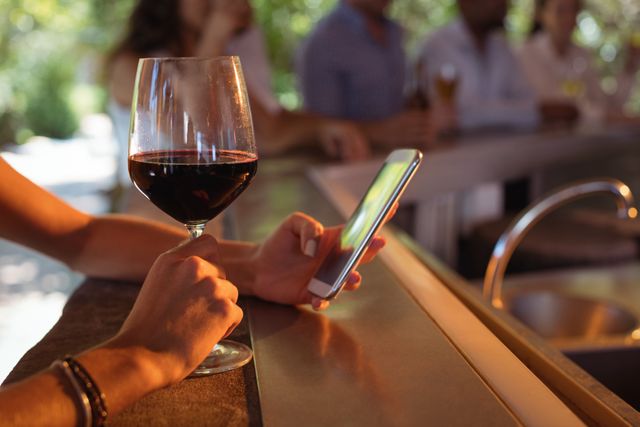 This image shows a woman holding a glass of red wine in one hand and using a mobile phone with the other at a restaurant bar. Ideal for use in articles or advertisements related to dining out, technology in social settings, or lifestyle and leisure activities.