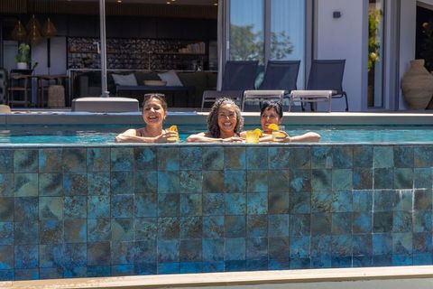 Diverse women leaning in tiled pool holding citrus drinks, laughing and relaxing at backyard