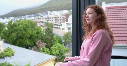 Redhead woman standing on balcony gazing over suburban rooftops and green hillside