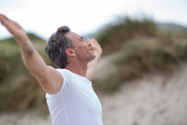 Mature man with grey hair doing meditation on the beach, with arms outstretched and eyes closed, expressing freedom and relaxation. Suitable for use in materials promoting mindfulness, healthy lifestyle, wellness, mental health practices, and outdoor activities. Perfect for websites, blogs, and advertisements focused on relaxation, peace, and nature.