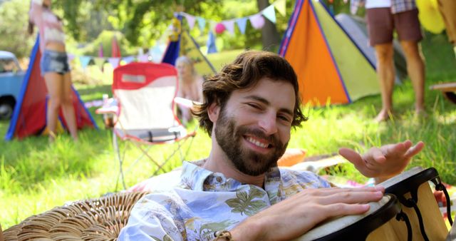 Man enjoying a summer festival amidst nature, playing bongo drum, surrounded by campers and festive decorations. Perfect for promotions related to music events, summer festivals, camping trips, relaxation and leisure activities in nature.