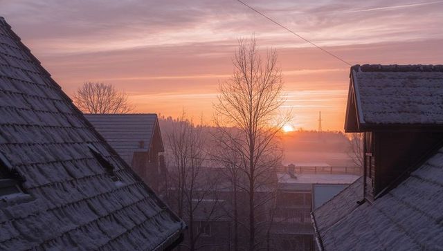 Sunrise framing snow-dusted roofs and misty fields with bare trees and pastel sky