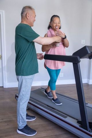 Senior asian couple exercising on home treadmill sharing water and supportive companionship