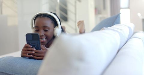 Smiling youth wearing white headphones on couch using smartphone, relaxed home moment