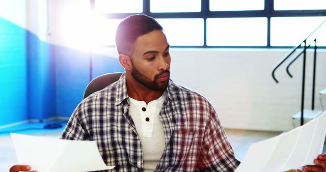 Middle-aged man in casual plaid shirt sits at desk intensively reviewing documents. Sunlight streams through the windows, contributing to a warm, focused atmosphere. Useful for themes of business, work, concentration, office life, and professional tasks.