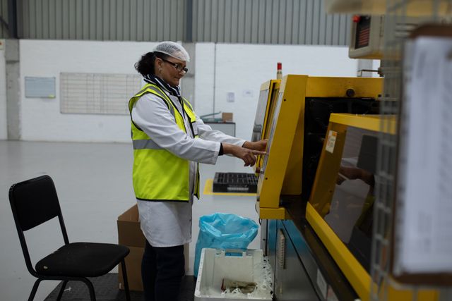 This image depicts a focused female worker in a factory warehouse, wearing a hair net, safety glasses, lab coat, and high visibility vest while operating a machine on the production line. Ideal for use in articles or advertisements related to manufacturing, industrial safety, workplace efficiency, and women in industry.