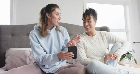 Friends sharing smartphone on bed, chatting and laughing in cozy sunlit bedroom