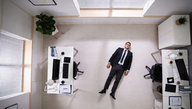 Overhead businessman lying on office floor between desks showing corporate burnout and pause