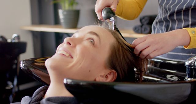 Woman receiving hair wash by stylist in a salon. Stylist's hands gently washing hair at shampoo station. Hair care, relaxation in salon. Ideal for promoting beauty services, hairdressing, self-care routines.