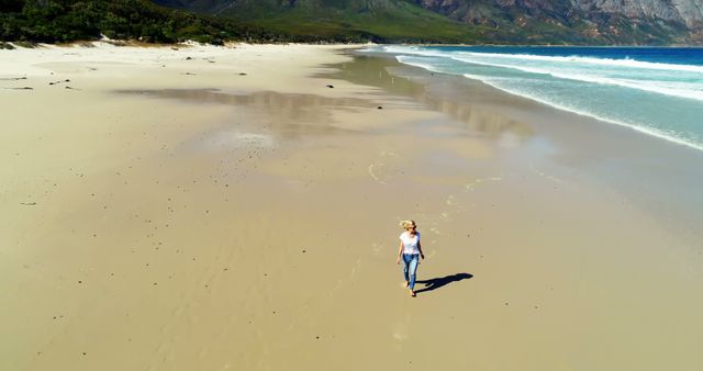 A woman is walking along a pristine and remote sandy beach. The scenery includes distant trees and mountains as well as gentle waves lapping at the shore. Perfect for concepts of travel, adventure, solitude, nature exploration, and relaxation.