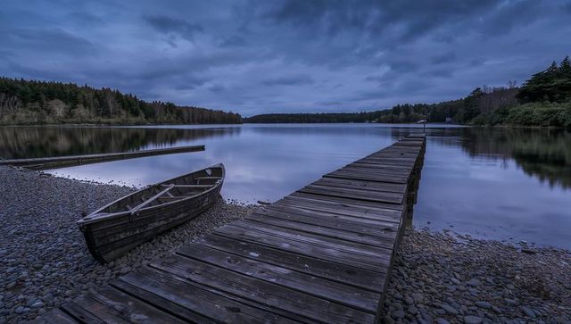Weathered wooden dock stretching over calm lake at dusk with rowboat on pebble shore