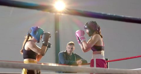 Female boxers sparring under spotlight while coach observing from ring corner with headgear