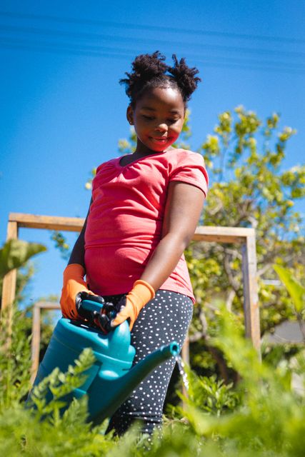 Young African American girl wearing orange gloves and a pink shirt, watering plants in a backyard garden under a clear blue sky. Ideal for use in educational materials about gardening, environmental awareness, and childhood activities. Perfect for lifestyle blogs, family-oriented content, and outdoor activity promotions.