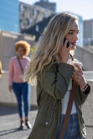 Young woman talking on phone holding coffee, urban commuter walking through sunlit plaza