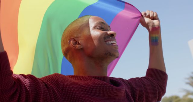 Happy african american male gay celebrating with pride flag outdoors in the sun. Pride, lgbtq, equality, rights and celebration, unaltered.