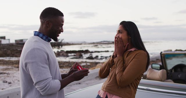 Man proposing to a happy, surprised woman by the seaside. They are next to a convertible car, with a picturesque ocean view in the background. Great for use in advertising, relationship articles, romance themes, and engagement announcements.