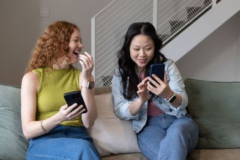 Friends laughing and sharing smartphones on sofa, wearing smartwatches and denim