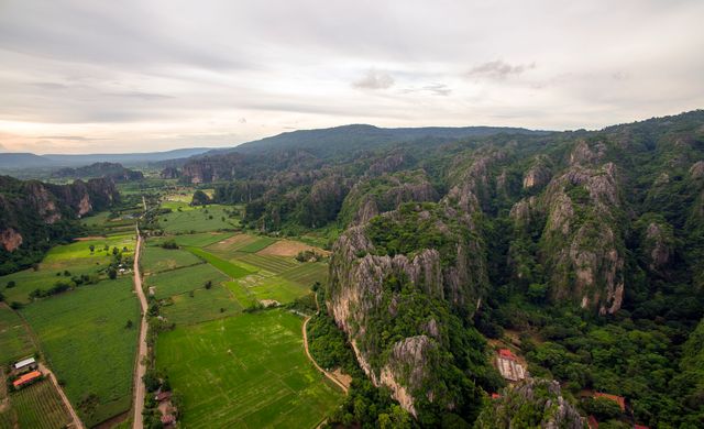 A breathtaking aerial view showcasing lush green valley surrounded by towering limestone mountains. Fields of green grass stretch across the horizon, interspersed with farms and small trees. Ideal for promoting tourism, eco-tourism, travel adventures, or nature conservation initiatives. Great for use in magazines, websites, travel brochures, or environmental awareness campaigns.