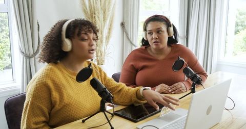 Two women recording podcast in sunlit home studio wearing headphones using laptop