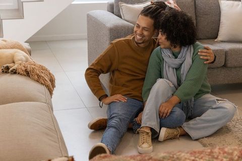 African american couple sitting on floor cuddling and watching dog on ottoman in living room
