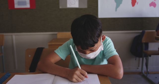 Diverse boy sitting at desk, wearing facemask and writing in notebook in elementary school class. School, learning, childhood, education, unaltered, health, hygiene, coronavirus.