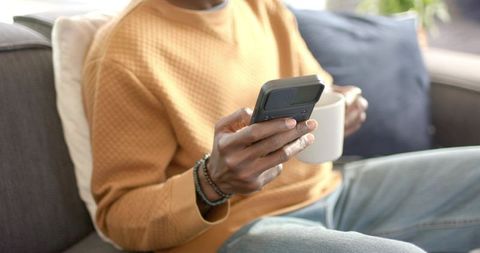 African American Man Relaxing on Sofa Checking Phone Holding Coffee Mug in Sunlit Home