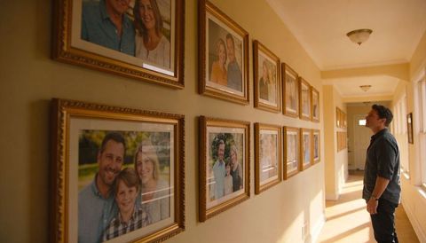 Man reflecting while viewing framed family portrait wall in warm sunlit home hallway