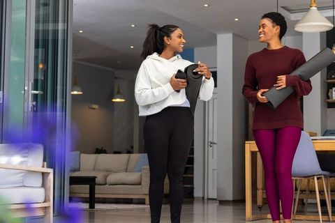 Diverse women holding yoga mats, chatting in modern living room, prepping home workout