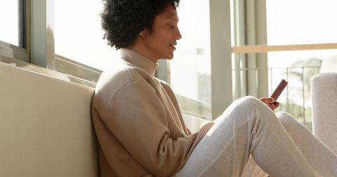 African american man sitting by window using smartphone in sunlit minimalist living room