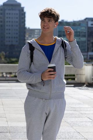 Teenage male standing in grey tracksuit holding coffee and adjusting earbud on urban plaza