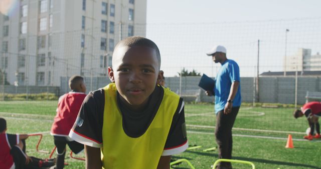 Boy smiling while training on a soccer field. Coach and other teammates in background focusing on drill routines. Ideal for promoting youth sports programs, teamwork, physical fitness, community activities, and athletic training camps.