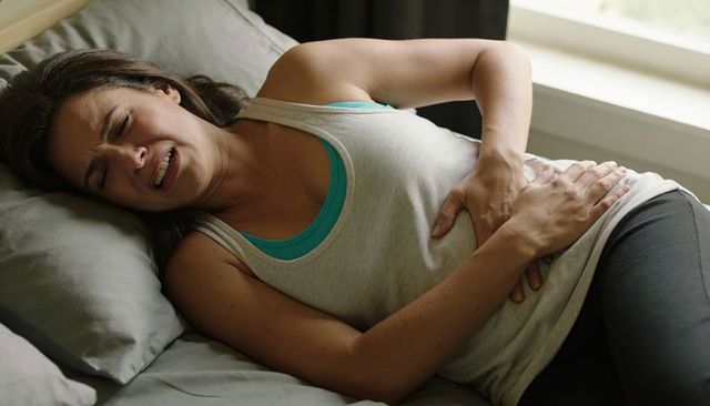 Young woman experiencing severe abdominal pain lying on bed holding stomach by window daylight